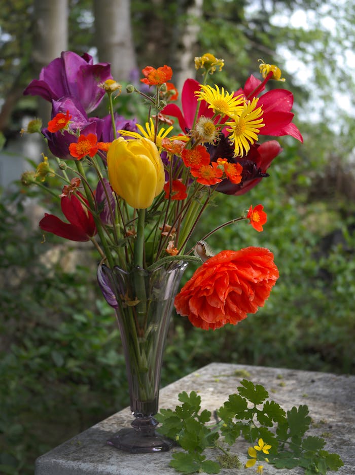 A colorful bouquet of spring flowers in a glass vase, set outdoors, showcasing nature's beauty.